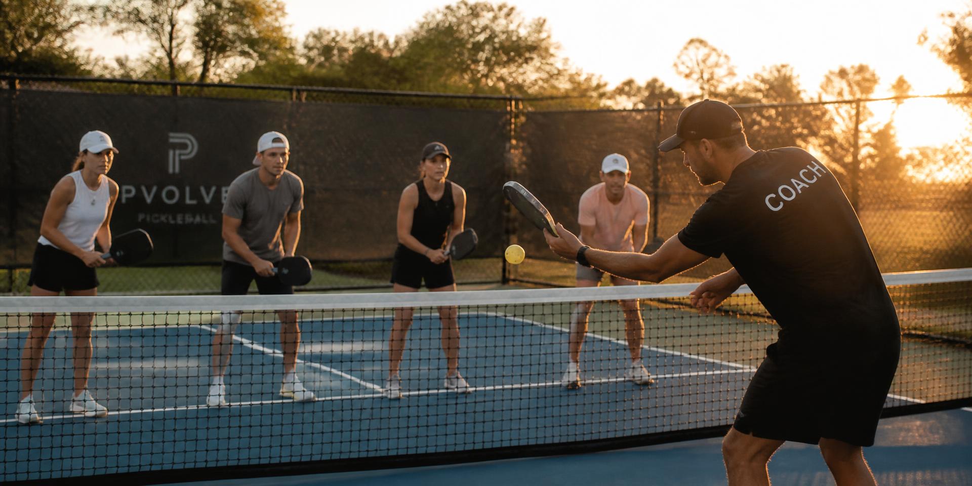 Coach teaching a group of players on an outdoor pickleball court at sunset